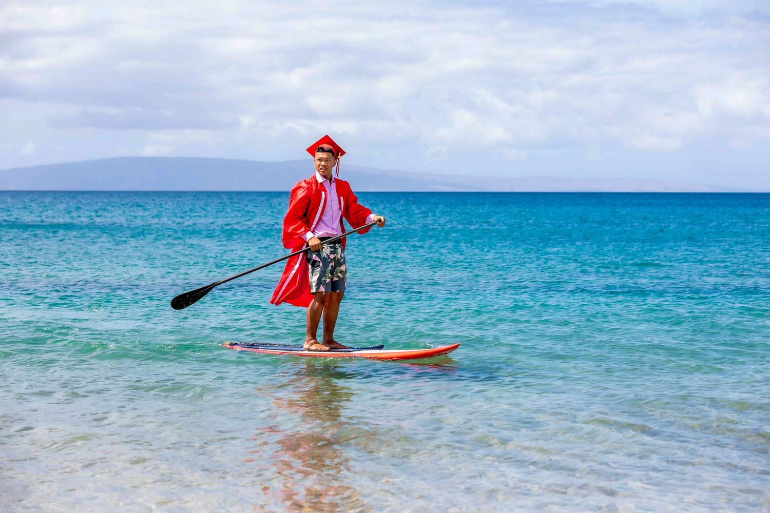 Person in a red pirate costume and hat stand-up paddleboarding on calm turquoise tropical water with distant mountains under a cloudy sky.