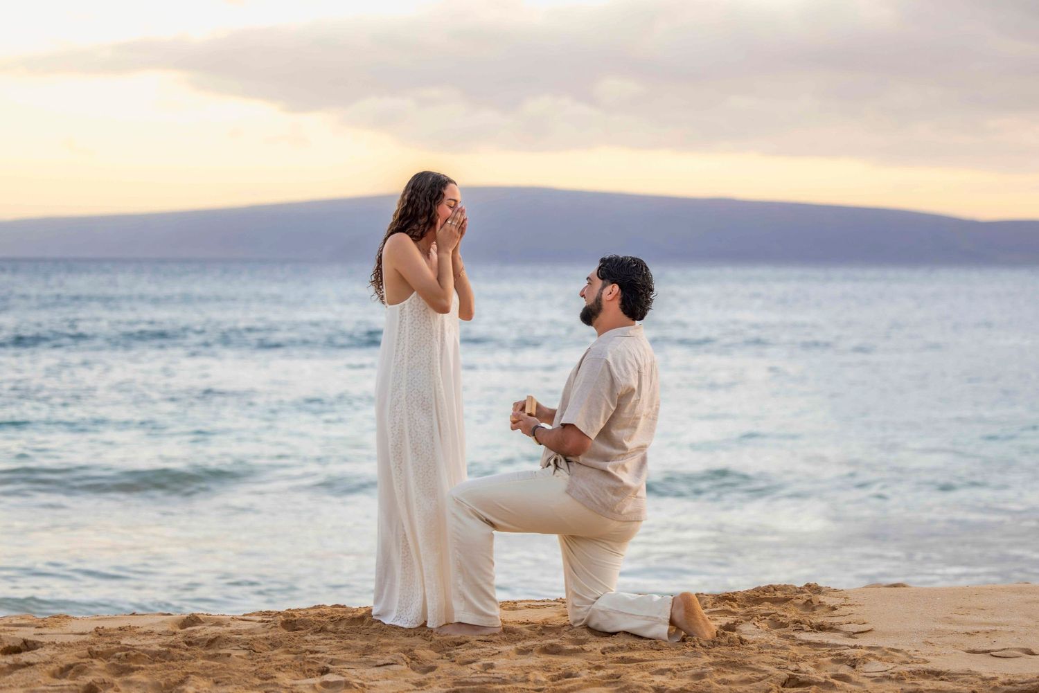 Man kneeling on a sandy beach at sunset proposing to a woman in a white dress who covers her face in surprise by the ocean. Makena