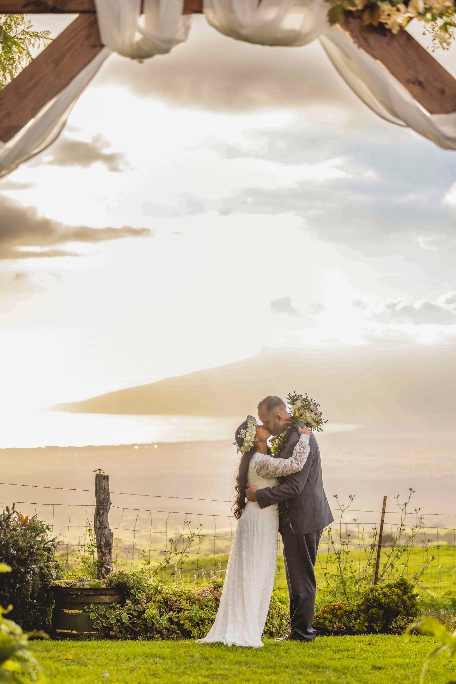 Bride in a white lace gown and groom in a gray suit kiss under a wooden wedding arch at sunset with an ocean and hills view. Kula