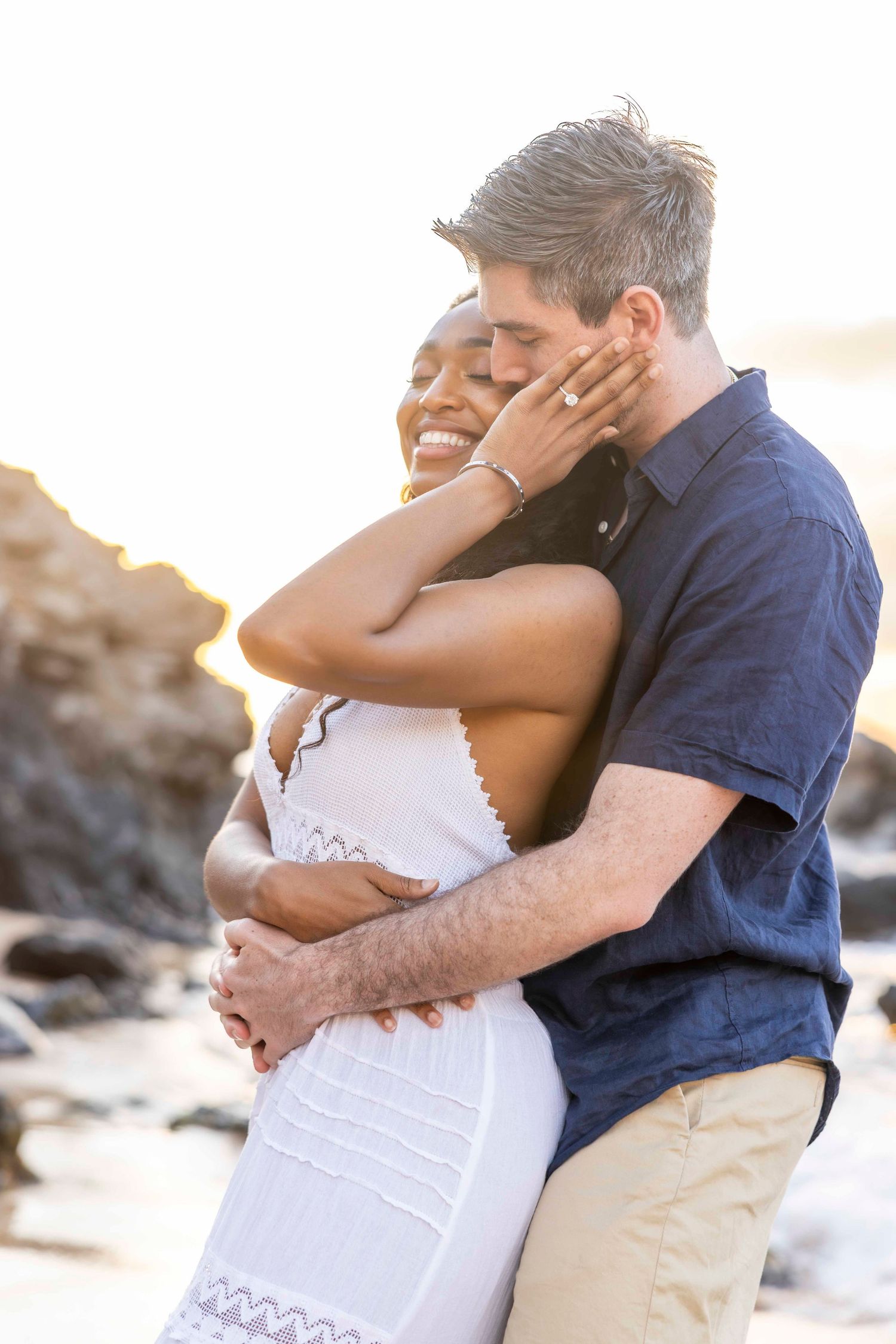 Smiling couple embracing on a sunlit rocky beach at sunset, she in a white dress touching his hand, he in a navy shirt holding her. Kapalua