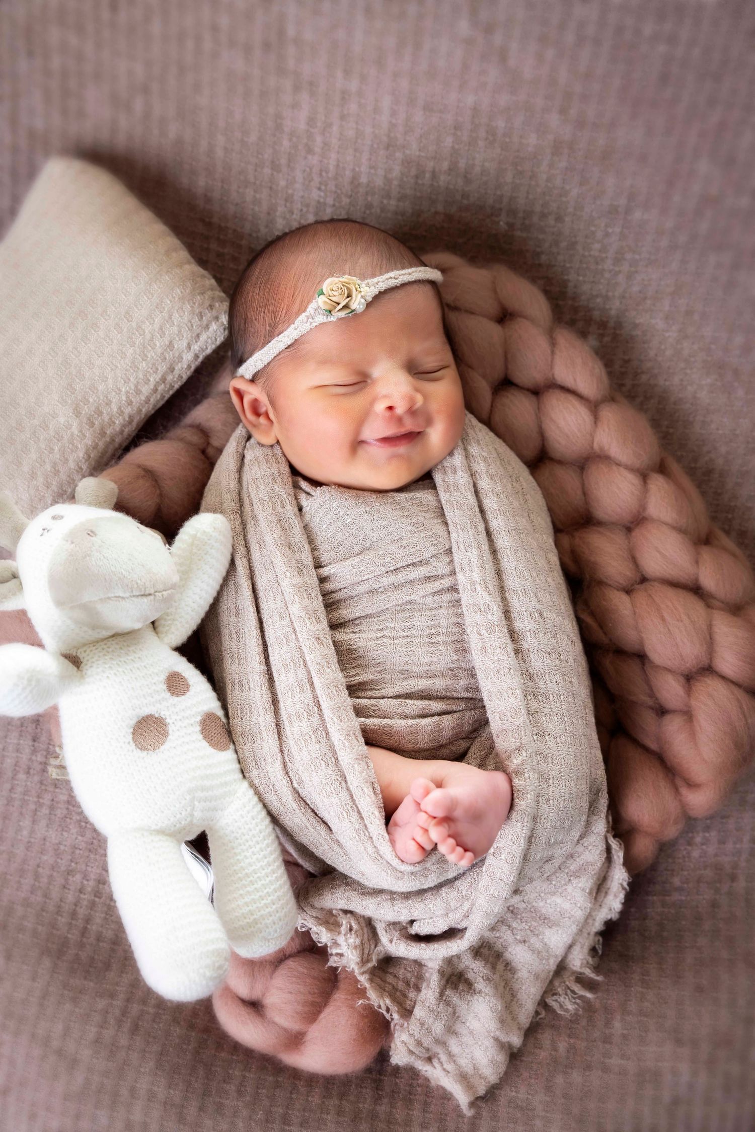 Sleeping newborn in a beige wrap and floral headband rests beside a plush giraffe toy on chunky knit blankets.