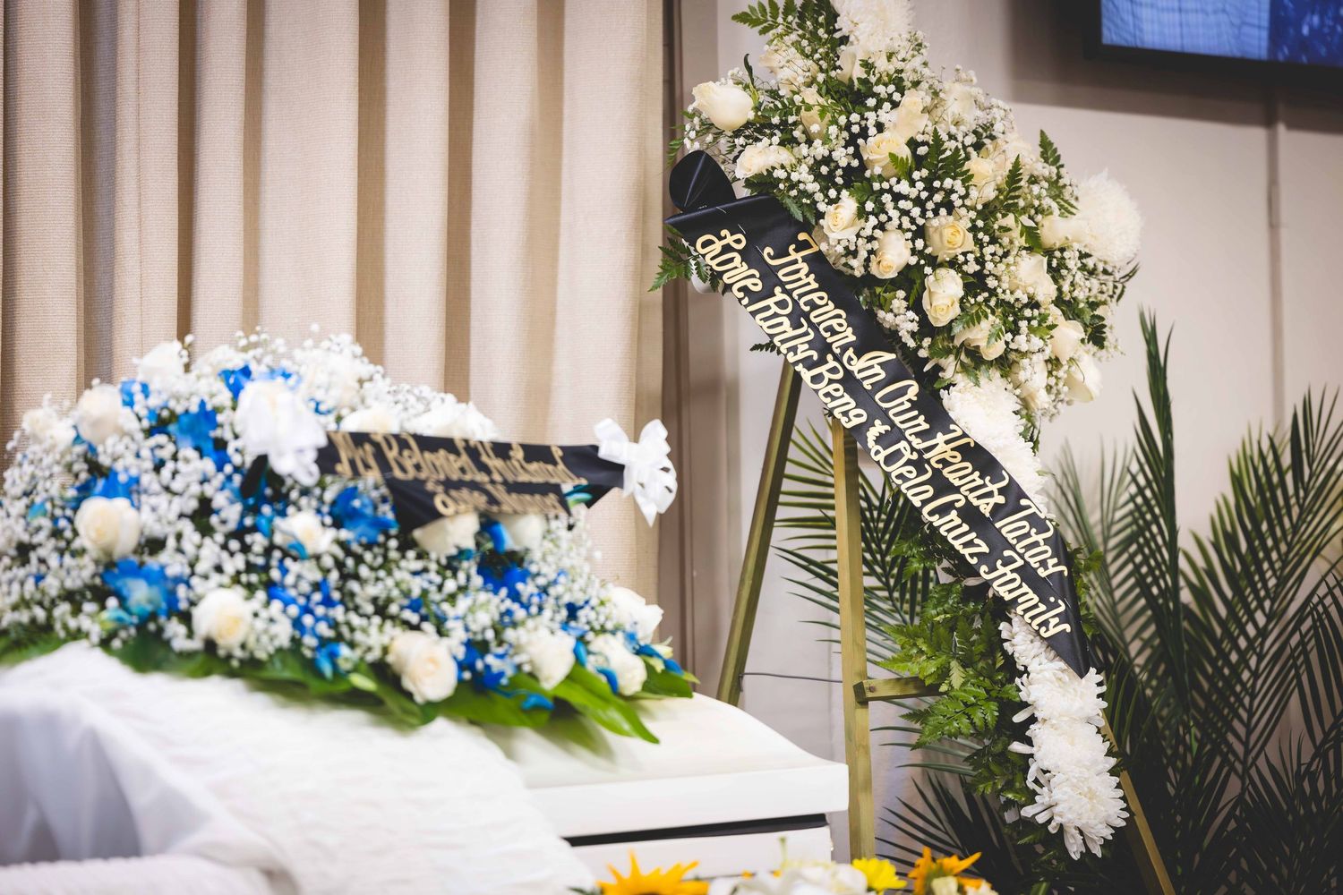 Blue and white funeral flower arrangement with roses on a casket, beside a standing wreath with a black ribbon sash and palm fronds.