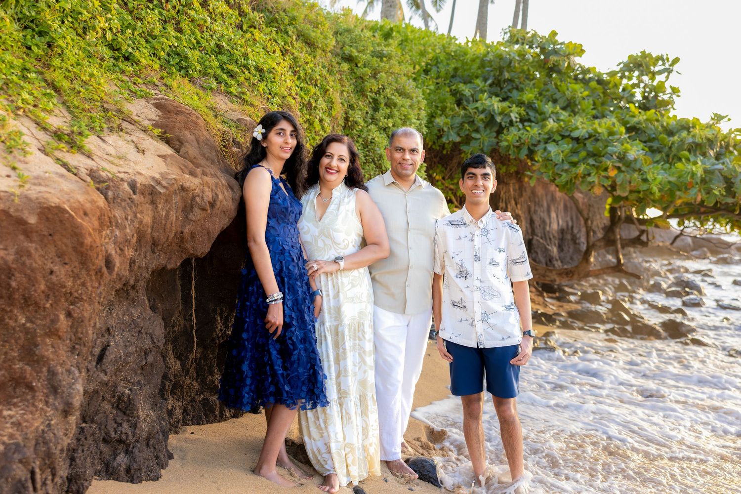 Family of four in coordinated blue and white outfits poses on a beach beside a rocky cliff and gentle waves in warm evening light.