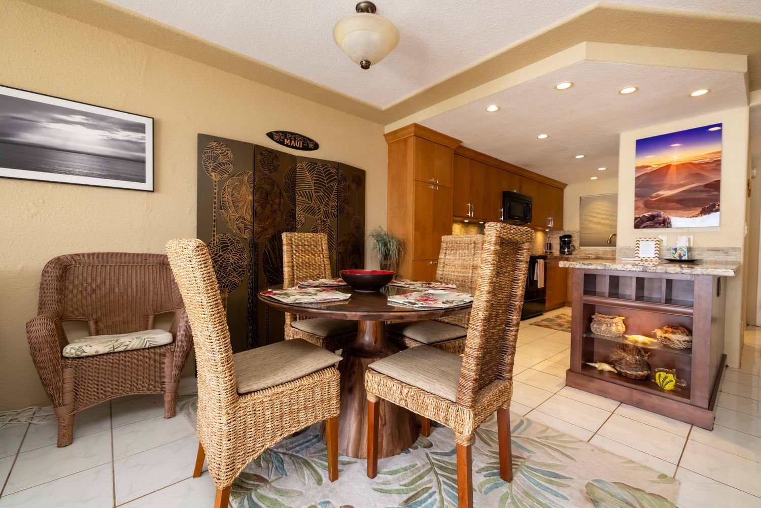 Bright dining area with a round wooden table, wicker chairs, tile floor, wood cabinets, and framed beach artwork in a vacation condo.