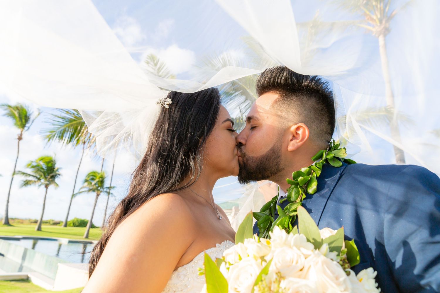 Bride and groom share a romantic kiss under a flowing white veil, with a bouquet and green leaf lei beside palm trees at a tropical wedding.