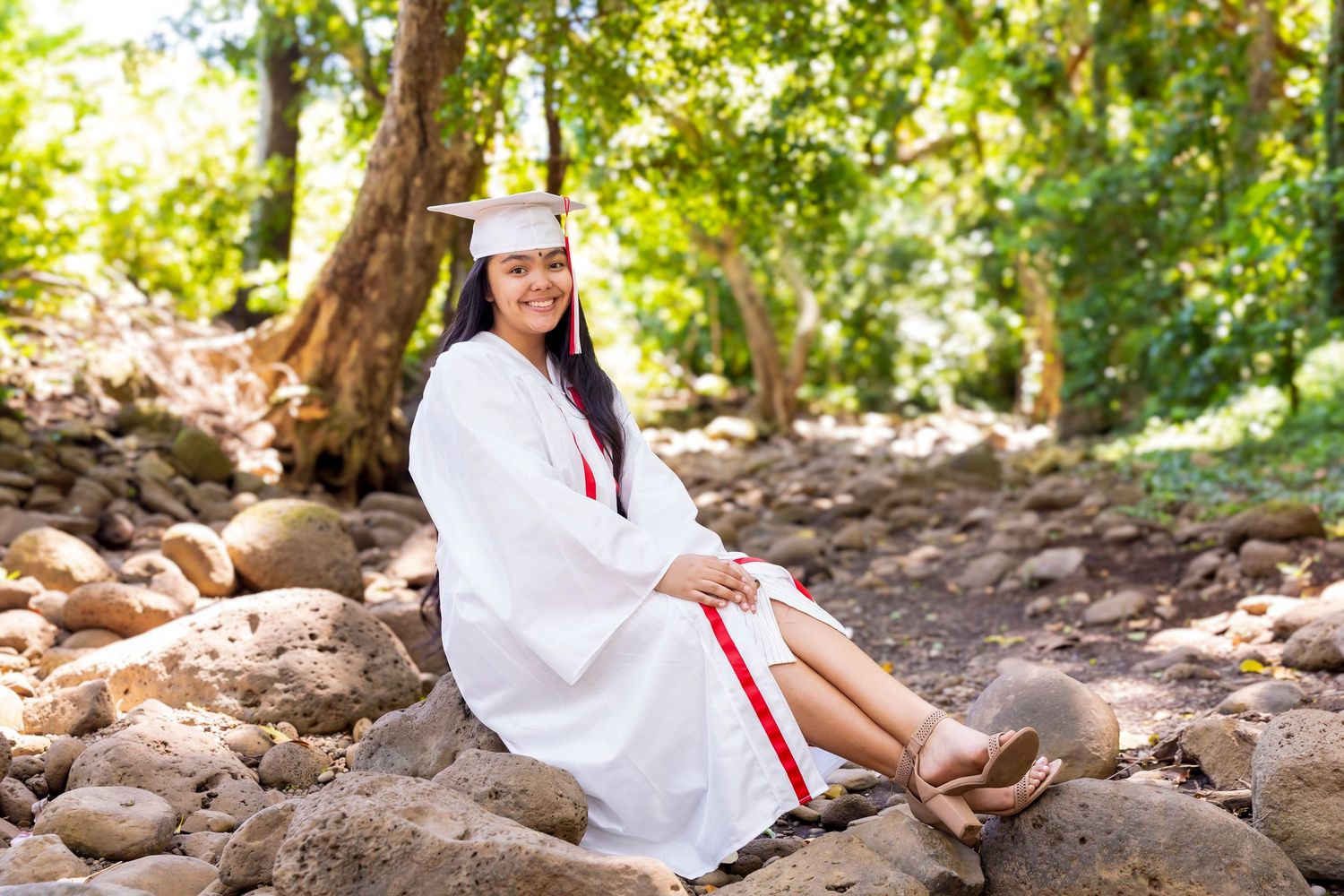 Graduate in a white cap and gown with a red stole sits on large rocks in a sunlit forest, posing for a outdoor graduation portrait. Honolua