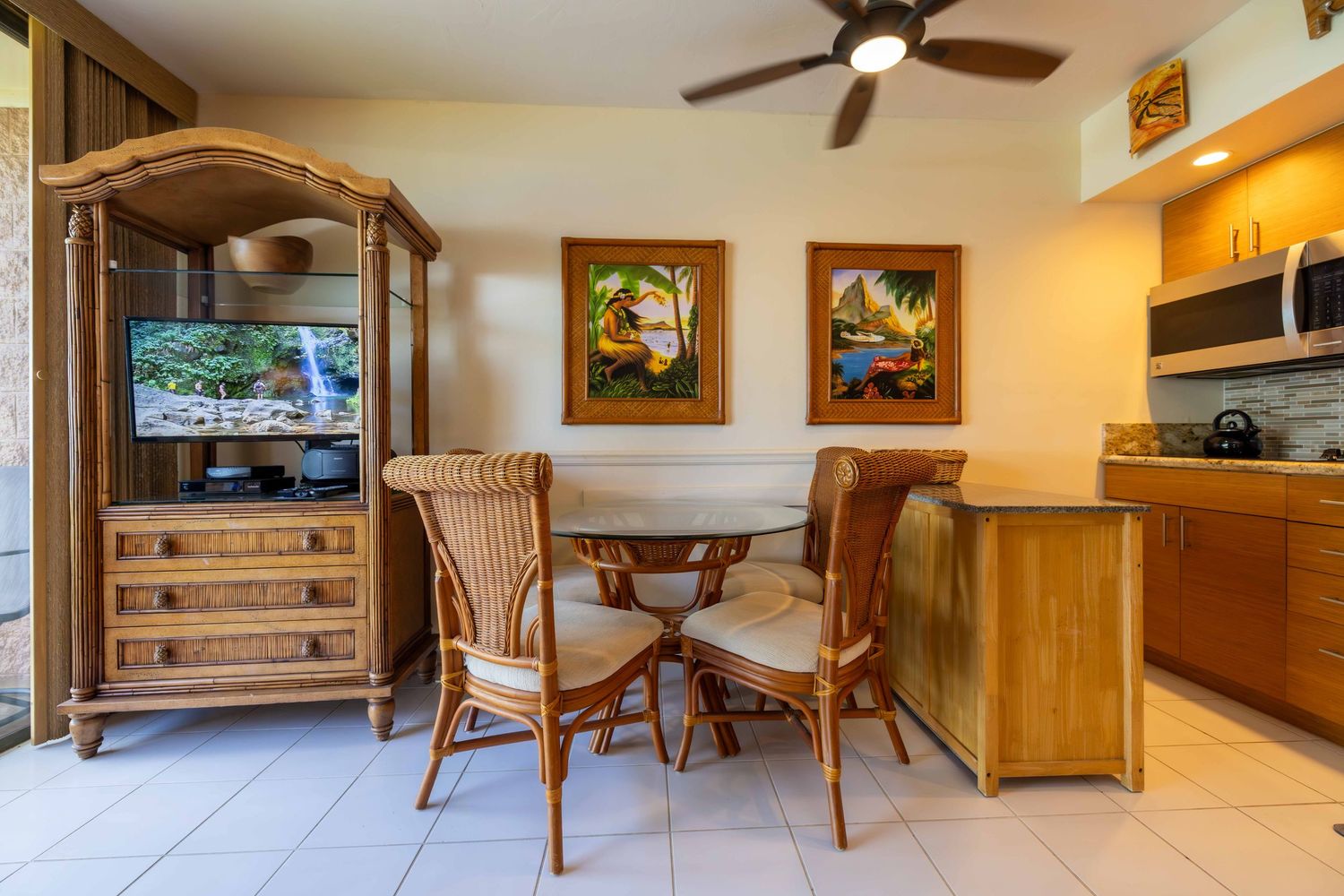 Bright vacation condo dining area with a round glass table, rattan chairs, tropical artwork, wood armoire with TV, and a kitchen counter.