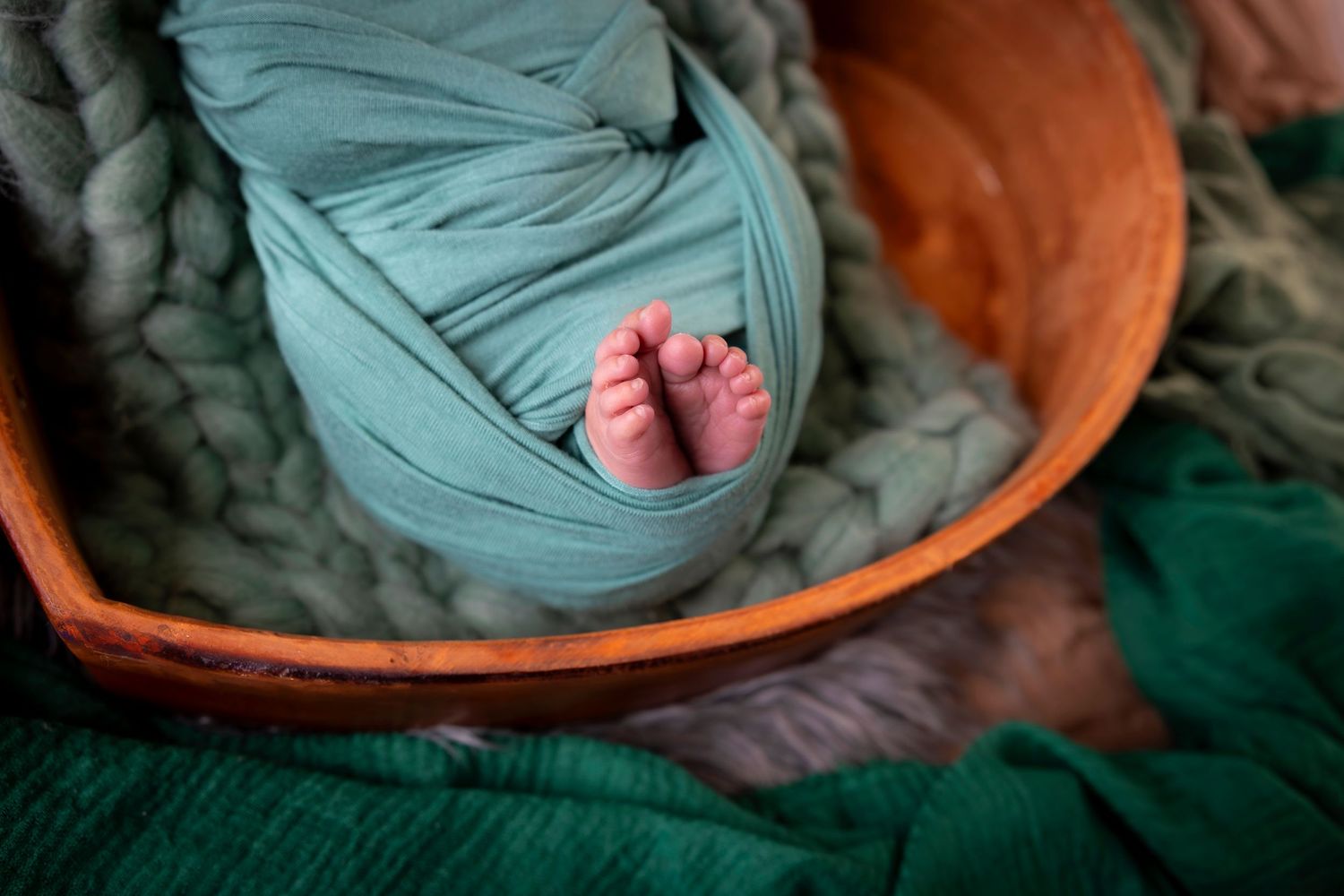 Close-up of tiny newborn feet peeking from a teal swaddle in a wooden bowl on chunky knit blankets.