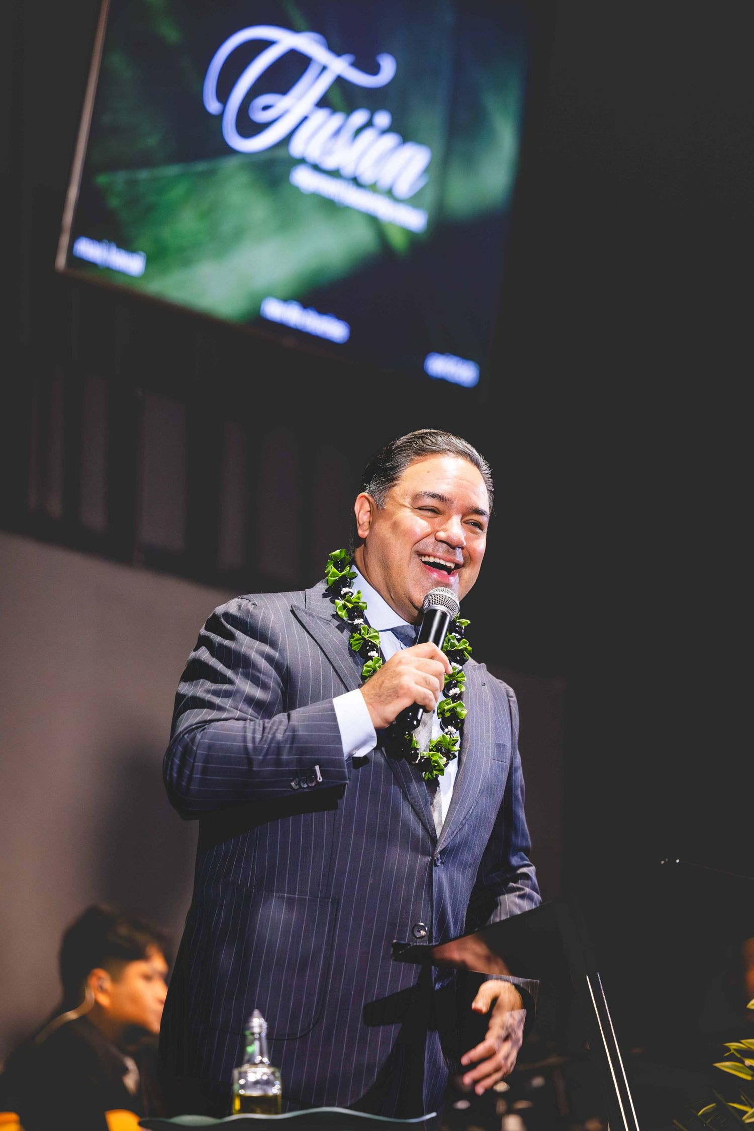 Smiling man in a pinstriped suit and green leaf lei speaks into a microphone on stage beneath a screen displaying the word Fusion.
