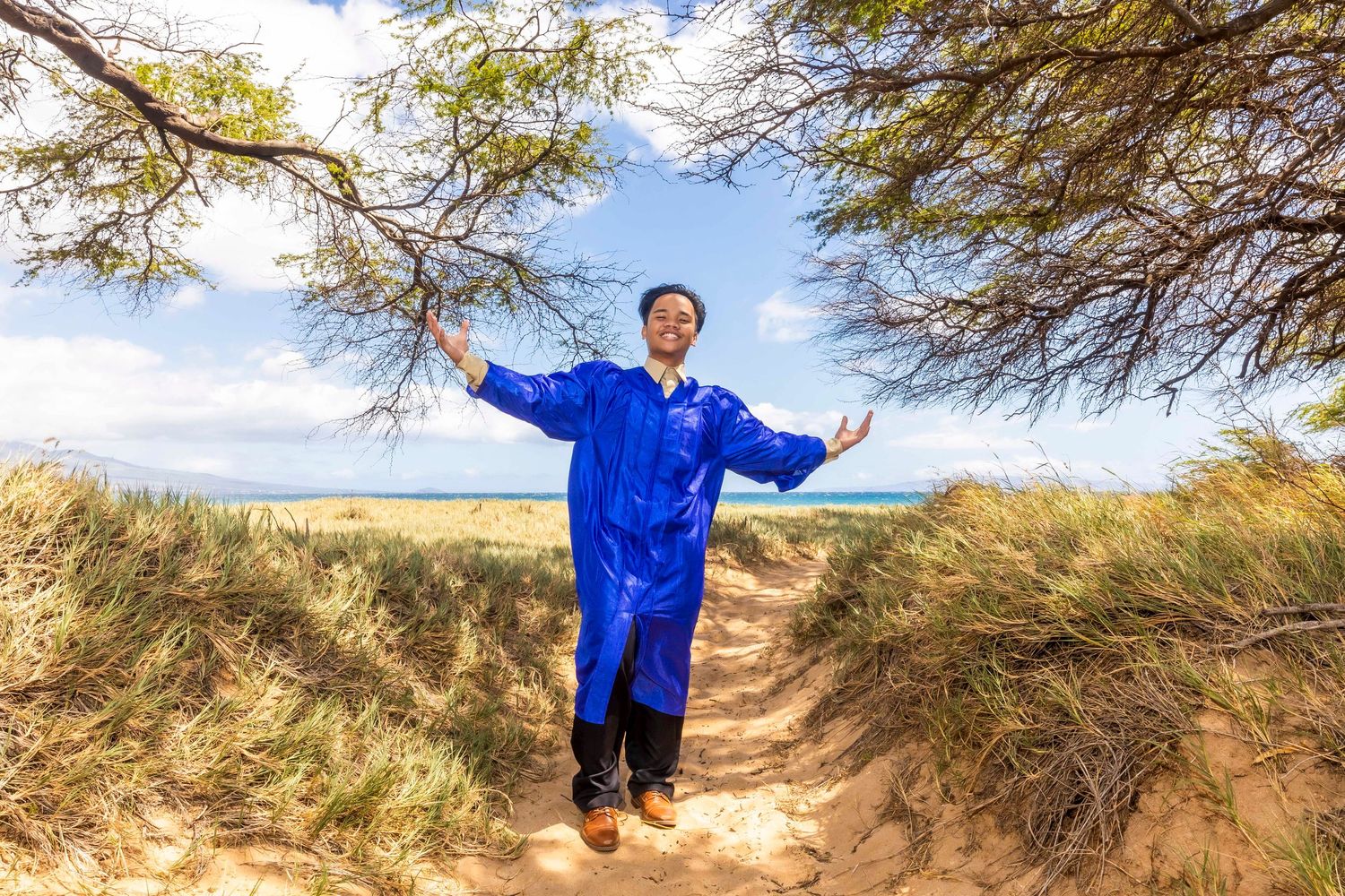 Person in a bright blue robe stands with arms outstretched on a sandy dune path, framed by tree branches and beach grass near the ocean.