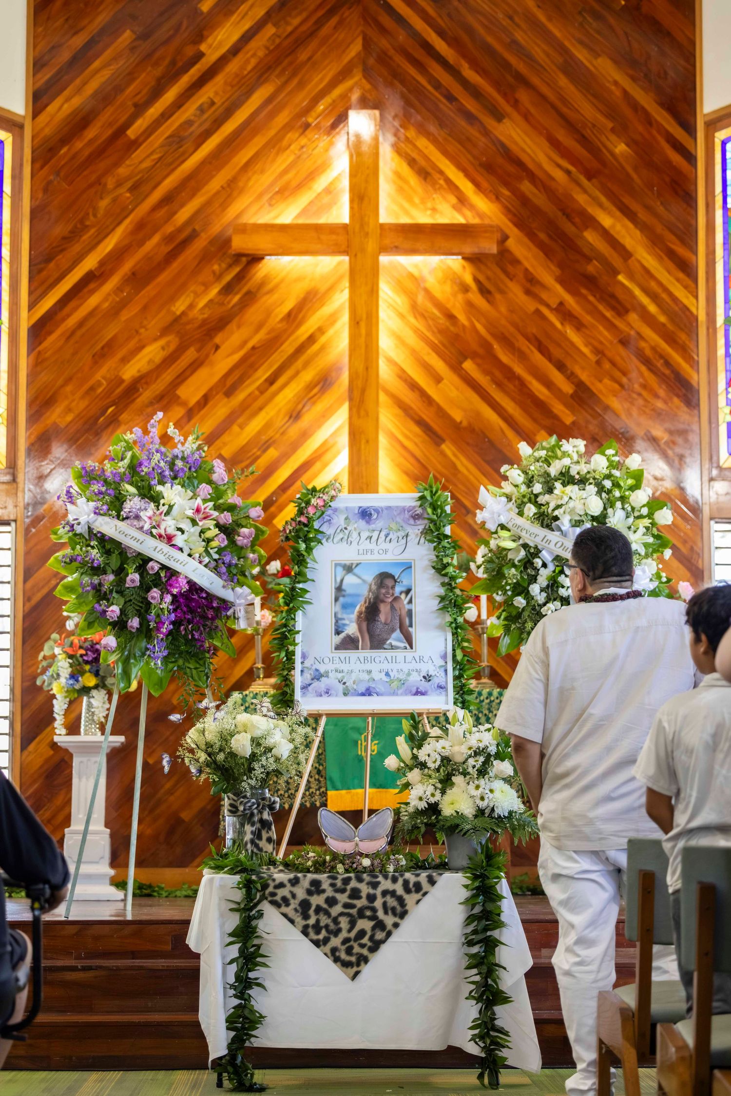 Memorial service in a church with a framed portrait, floral wreaths and a lei-draped table beneath a large wooden cross, as mourners attend.