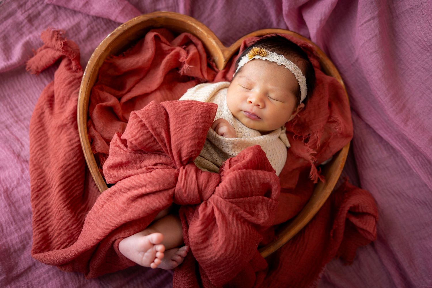 Newborn girl in a floral headband sleeps in a wooden bowl nest wrapped in rust red muslin on mauve fabric.