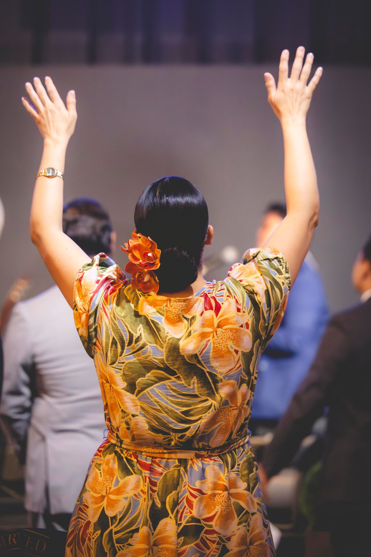 Woman in a colorful floral dress with an orange flower in her hair raises both hands in worship during a church service.