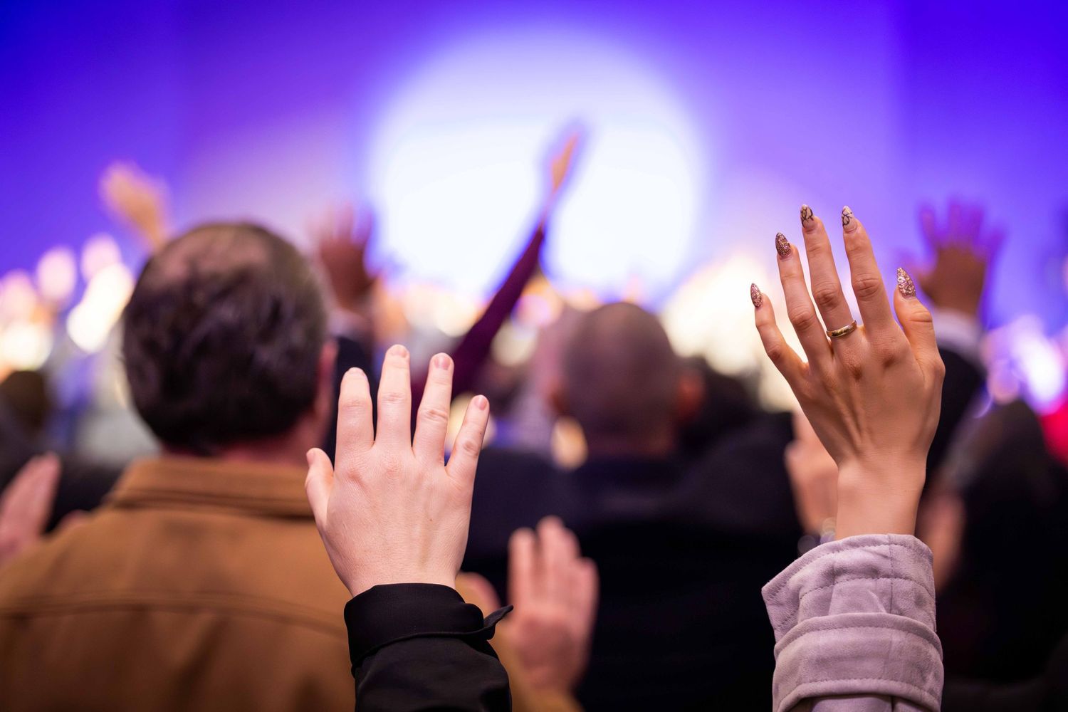 Worshippers raise their hands in prayer during a lively church service, with a glowing purple and blue stage backdrop blurred behind them.