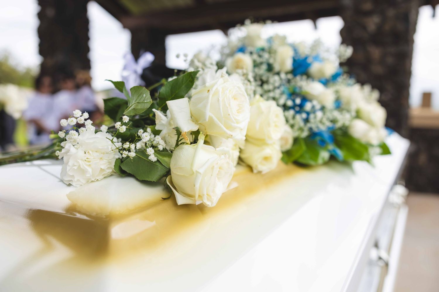 White roses, baby's breath and blue accent flowers arranged on top of a white casket during a funeral service.