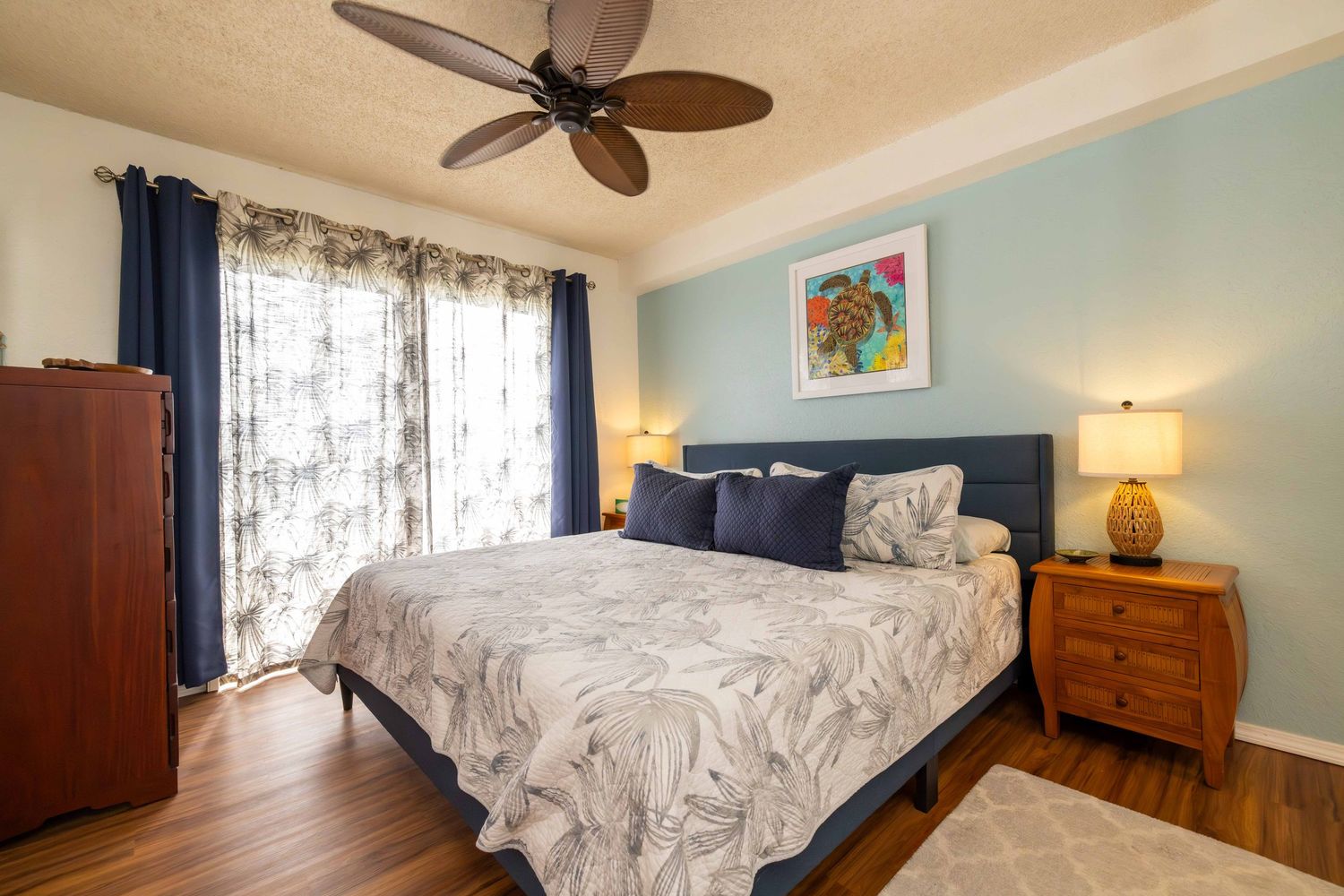 Cozy bedroom with a queen bed, blue accent wall, ceiling fan, wooden nightstand, tall armoire and sunny window with curtains.