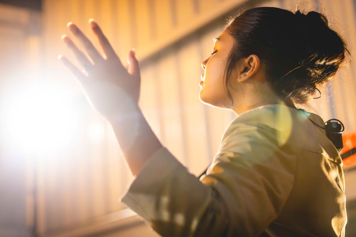 Woman in profile with eyes closed and hand raised in prayer, bathed in warm golden sunlight streaming through a window.