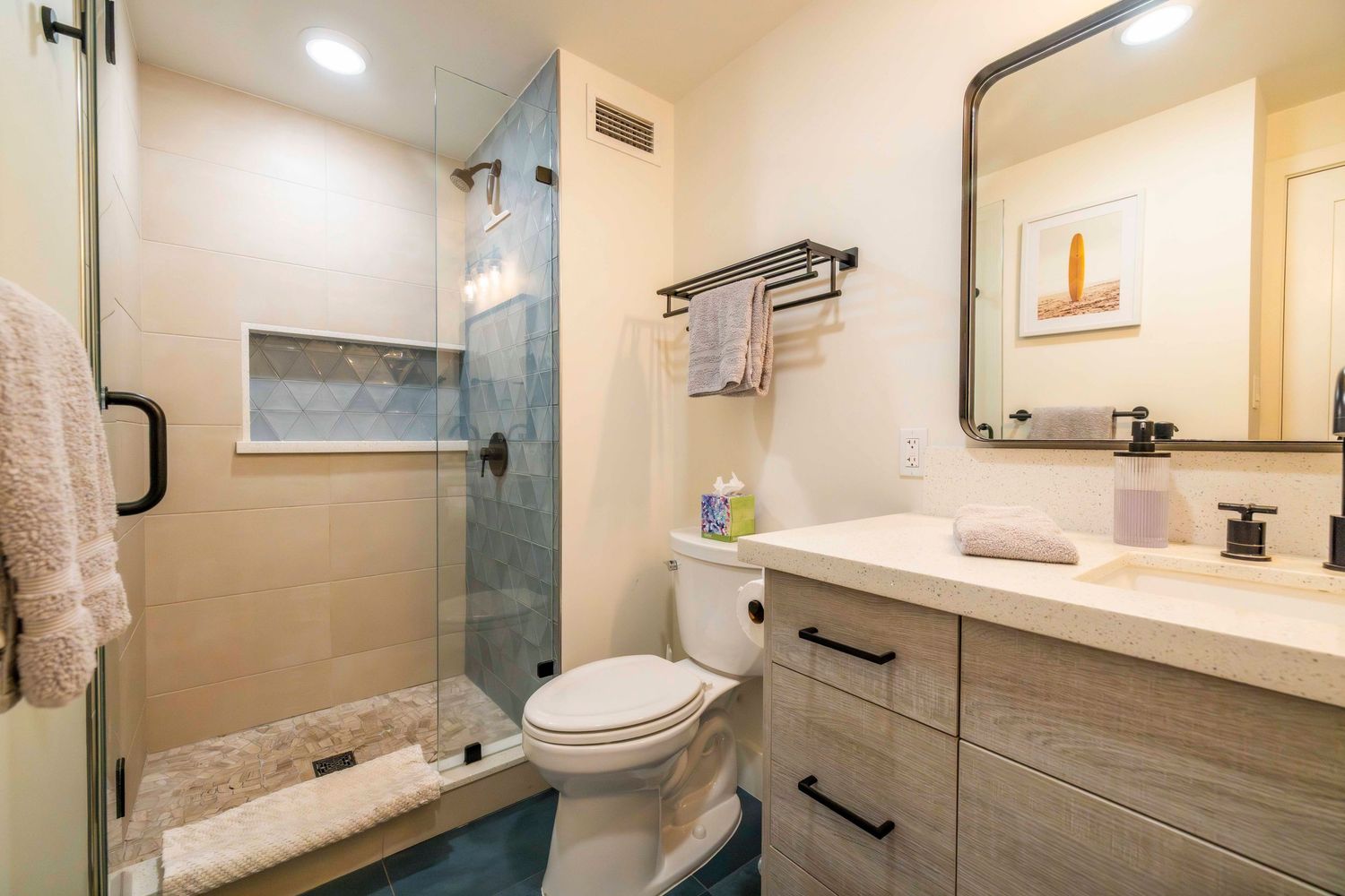Modern bathroom with a glass-enclosed tile shower, white toilet, and light wood vanity with black handles beneath a framed mirror.