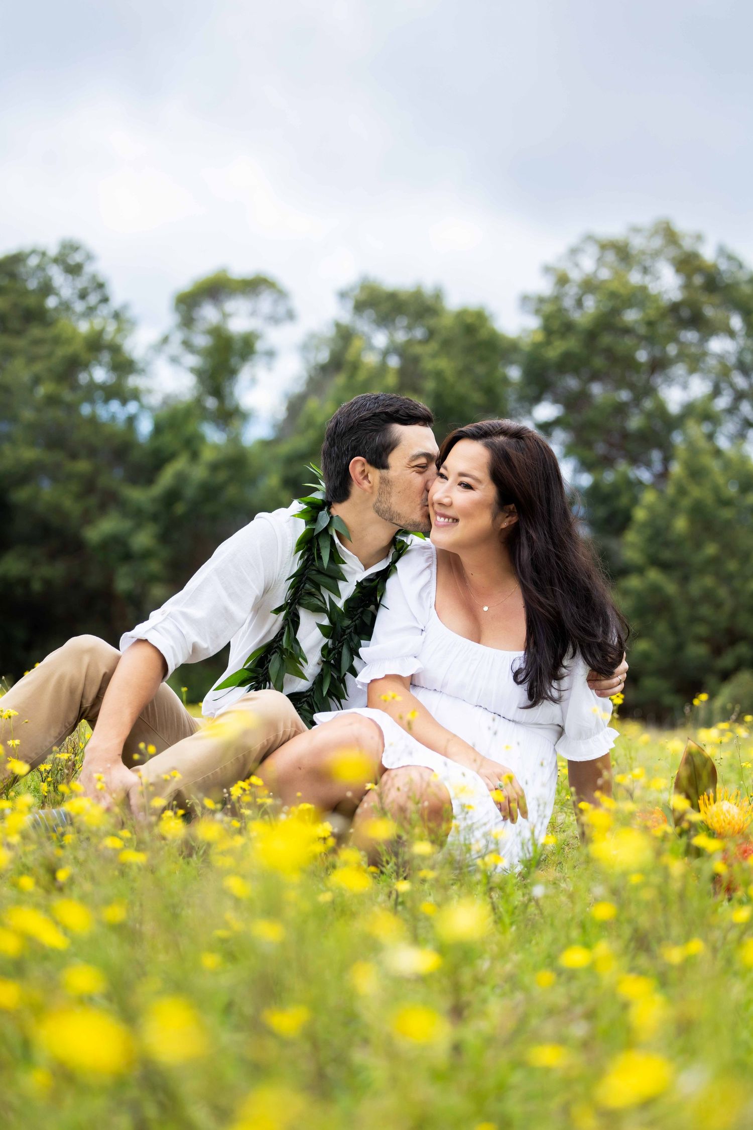 Couple sitting in a field of yellow wildflowers as the man wearing a maile lei kisses the smiling woman's cheek. Poli Poli