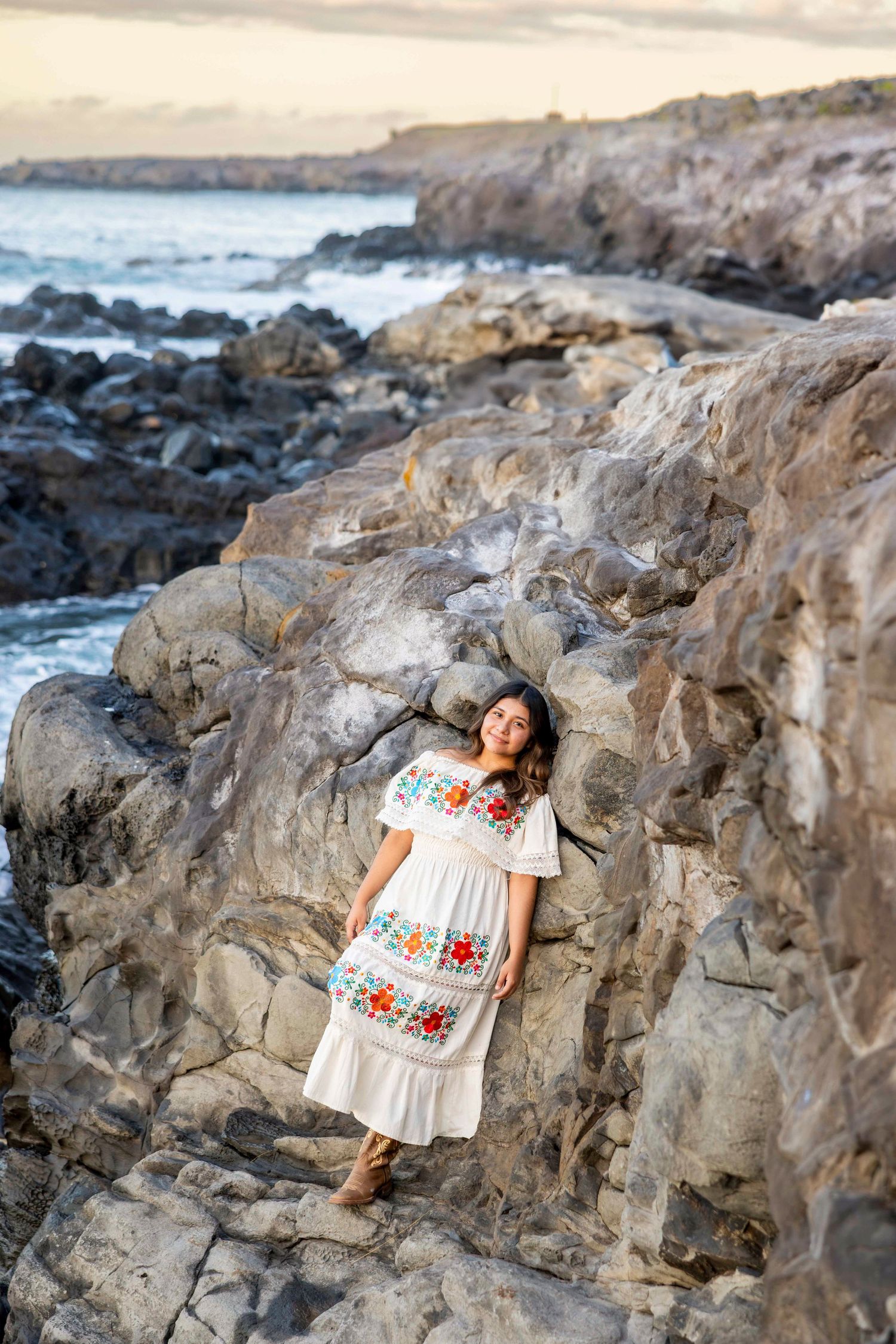Woman in a white embroidered floral dress leaning against rugged volcanic rocks by the ocean at sunset along a rocky coastline. Paia