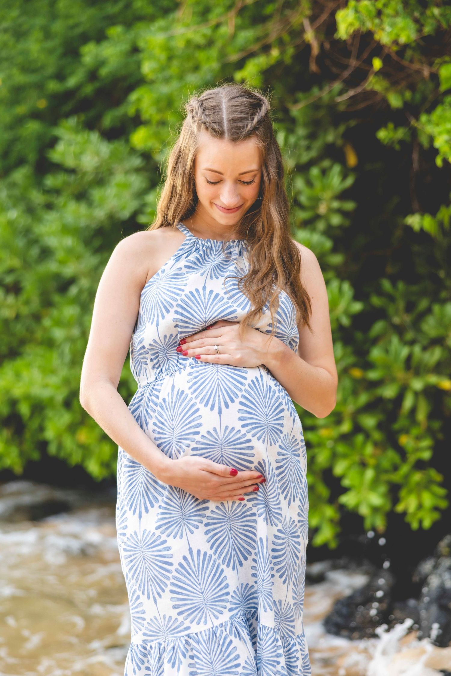 Smiling pregnant woman in a blue and white leaf-print dress cradles her baby bump beside lush green foliage and a rocky shoreline. Makena