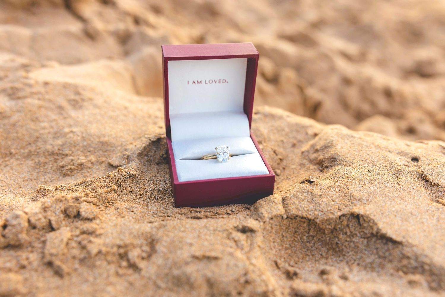 Open red ring box with a sparkling diamond engagement ring resting in golden beach sand, with the words I am loved inside the lid. Wailea