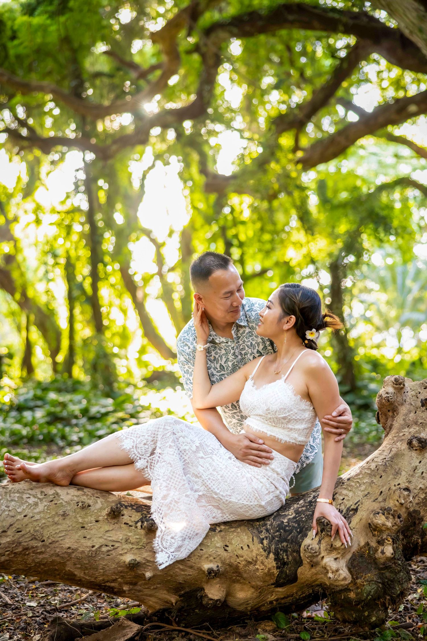 Romantic couple portrait of a woman in a white lace outfit sitting on a large tree branch, embracing a man under a sunlit green canopy.