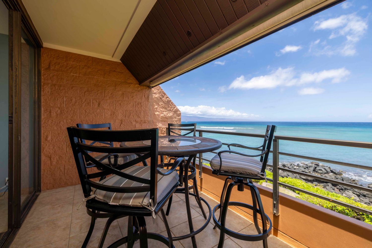 Oceanfront condo balcony with a bistro table and cushioned chairs overlooking a rocky shoreline and blue ocean under a partly cloudy sky.