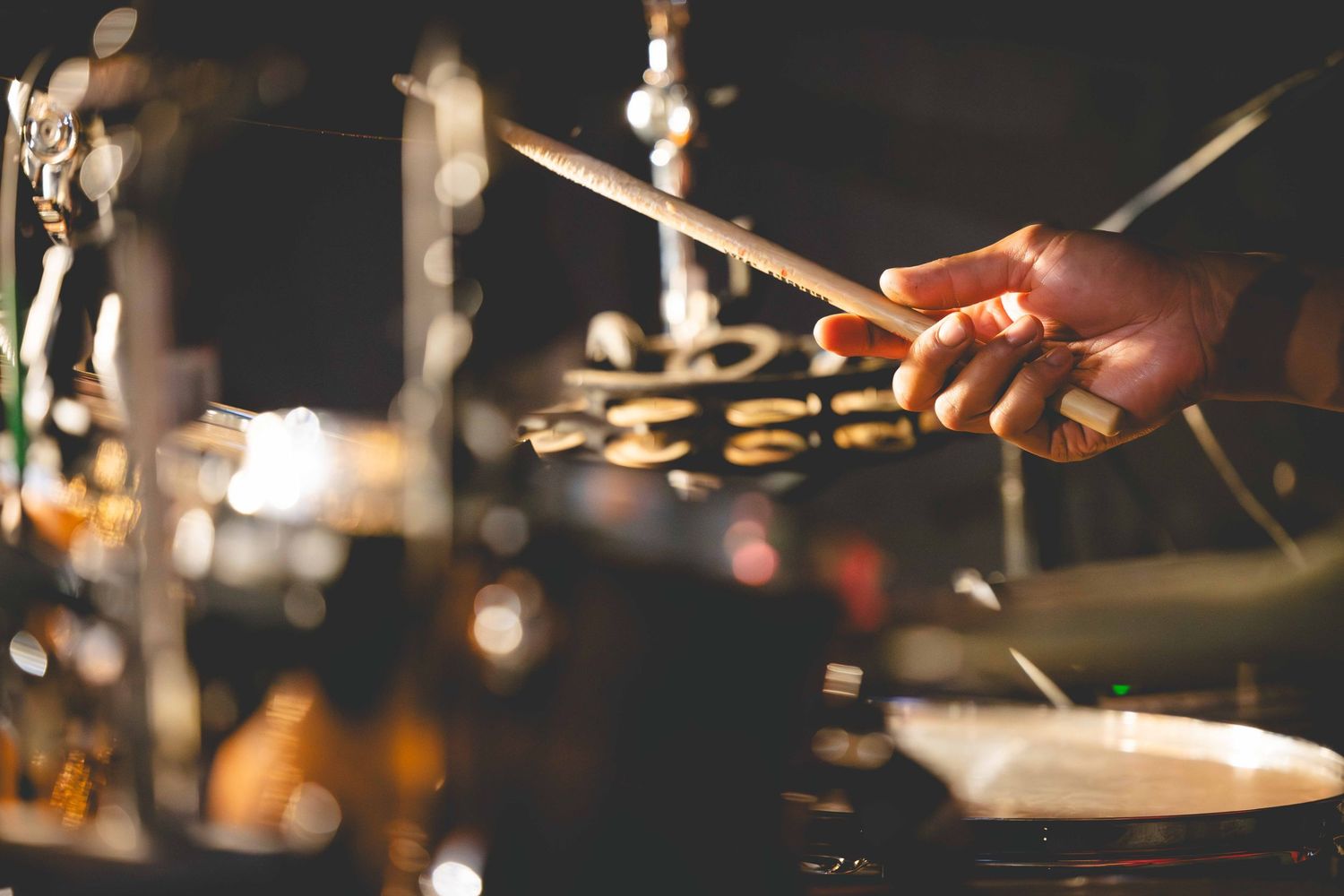 A drummer's hand holds a drumstick over a cymbal and hi-hat in a dimly lit stage setting with warm glowing lights.