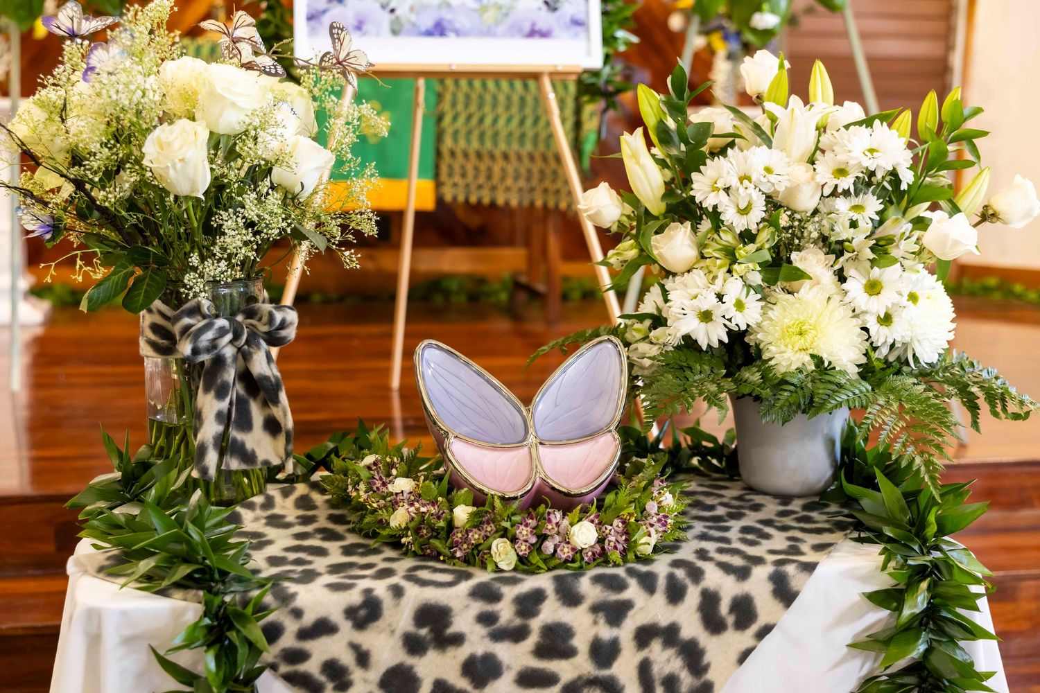 Pink and lavender butterfly-shaped urn surrounded by greenery and white flower arrangements on a leopard print cloth at a memorial service.