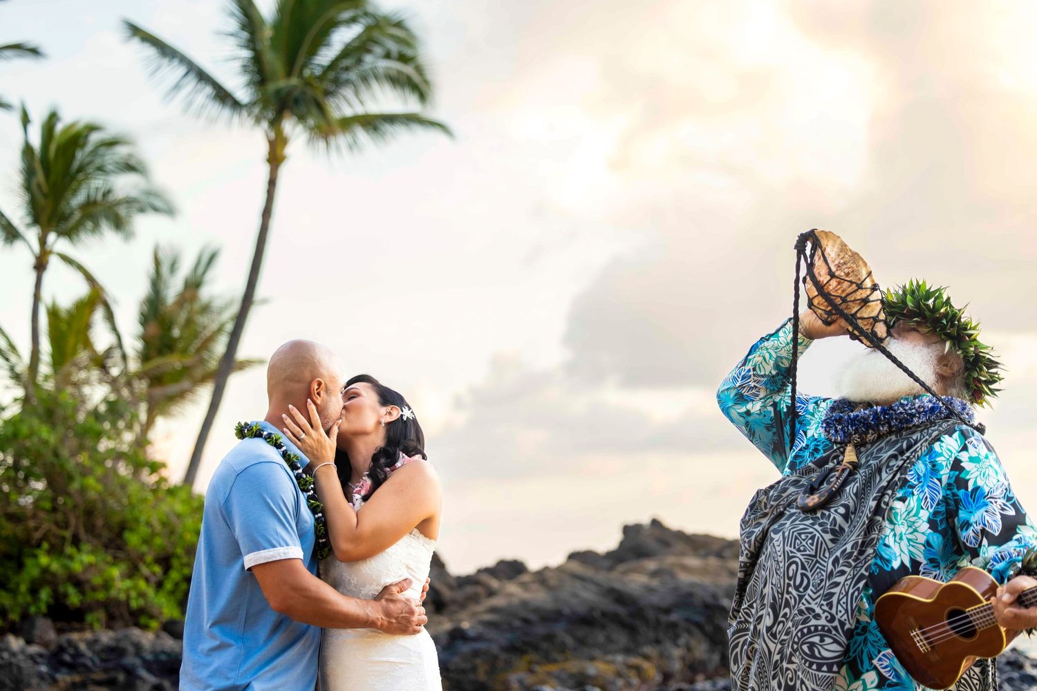 Couple kisses during a Hawaiian beach wedding ceremony as an officiant in a haku lei crown plays ukulele beside palm trees and lava rocks.