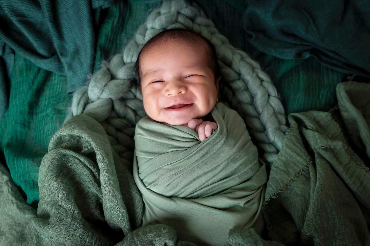 Smiling newborn baby swaddled in a sage green wrap lying on soft green blankets in a joyful newborn photo.
