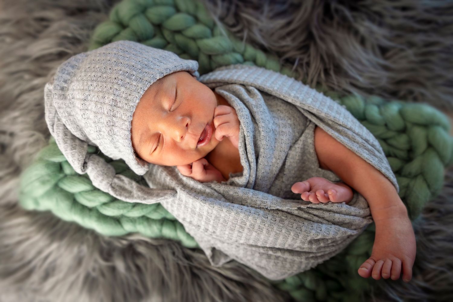 Newborn baby sleeping in a gray knit hat and wrap on a green chunky knit blanket and gray fur rug for a newborn photo shoot.
