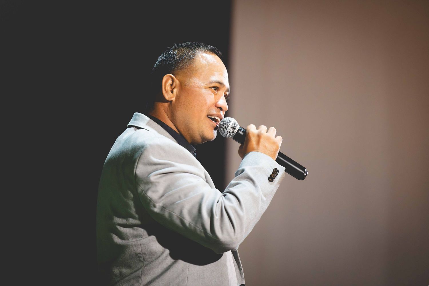 Man in a gray blazer smiles while speaking into a handheld microphone on a dimly lit stage.