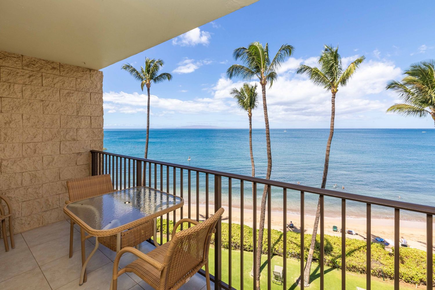 Oceanfront condo balcony with a patio table and chairs overlooking palm trees, a sandy beach, and the blue Pacific Ocean.
