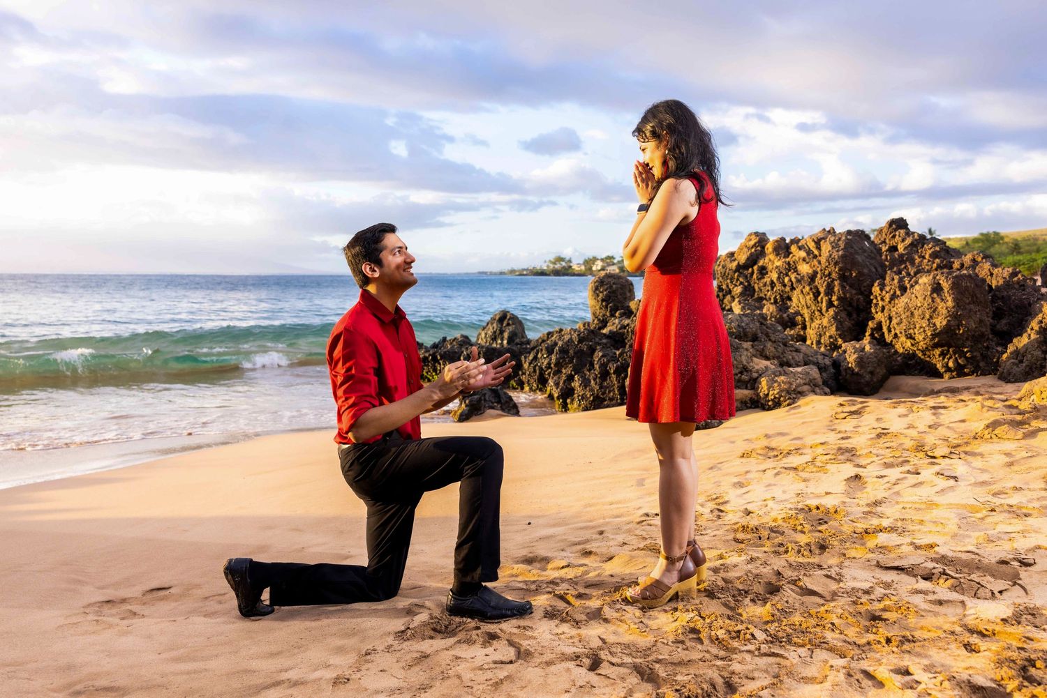 Man in red shirt kneels on one knee proposing to a woman in a red dress, covering her mouth in surprise, on a sunny beach with ocean waves.