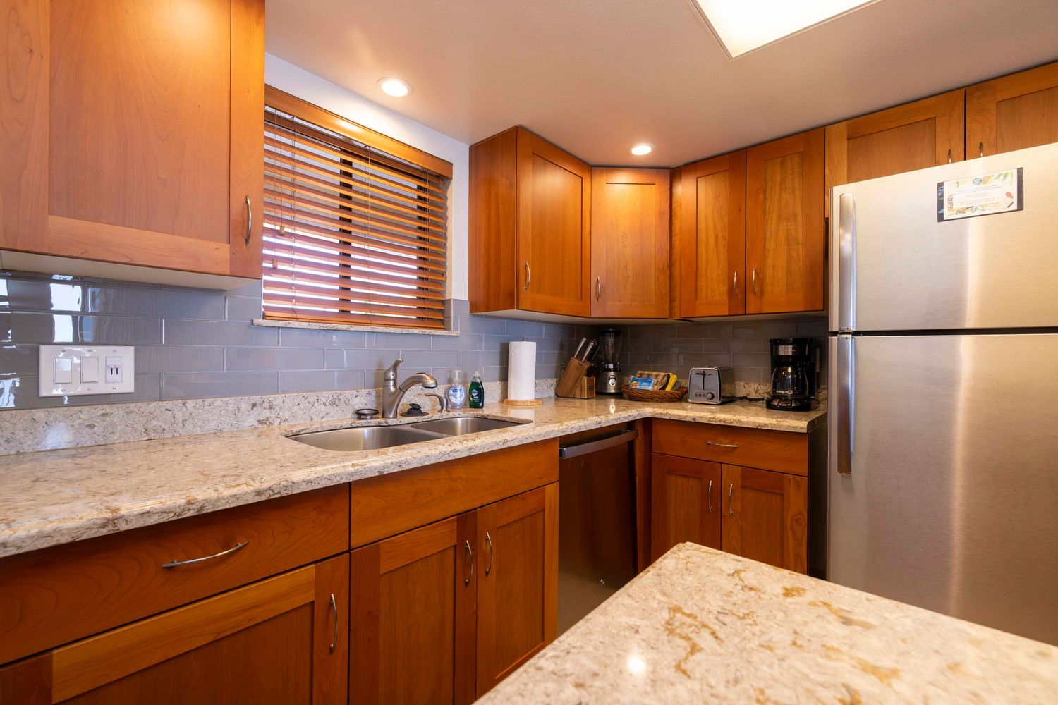Cozy kitchen with warm wood cabinets, granite countertops, stainless steel refrigerator, double sink, and window with wooden blinds.