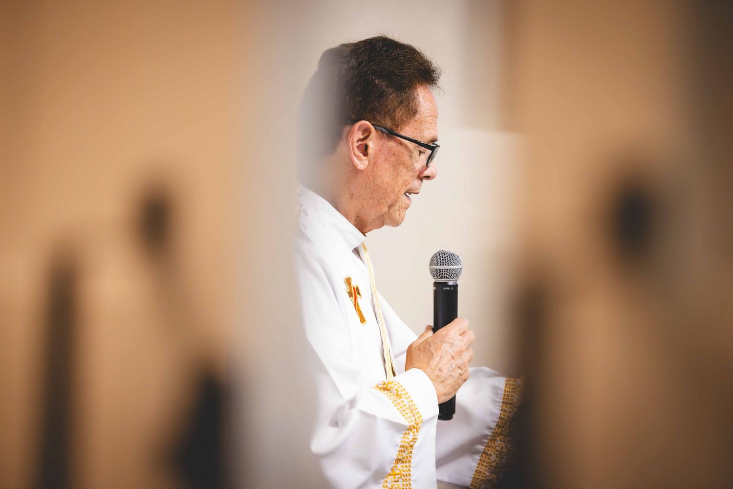 Priest in white vestments with gold trim and a cross speaks into a microphone during a church service, framed by softly blurred foreground.