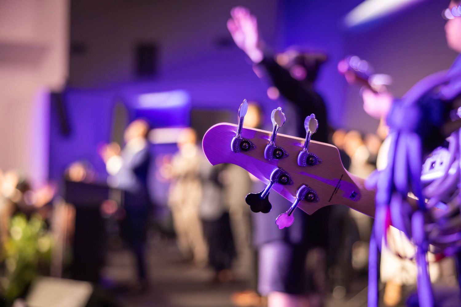 Close-up of a purple bass guitar headstock with tuning pegs, with blurred musicians and an audience on stage in the background.