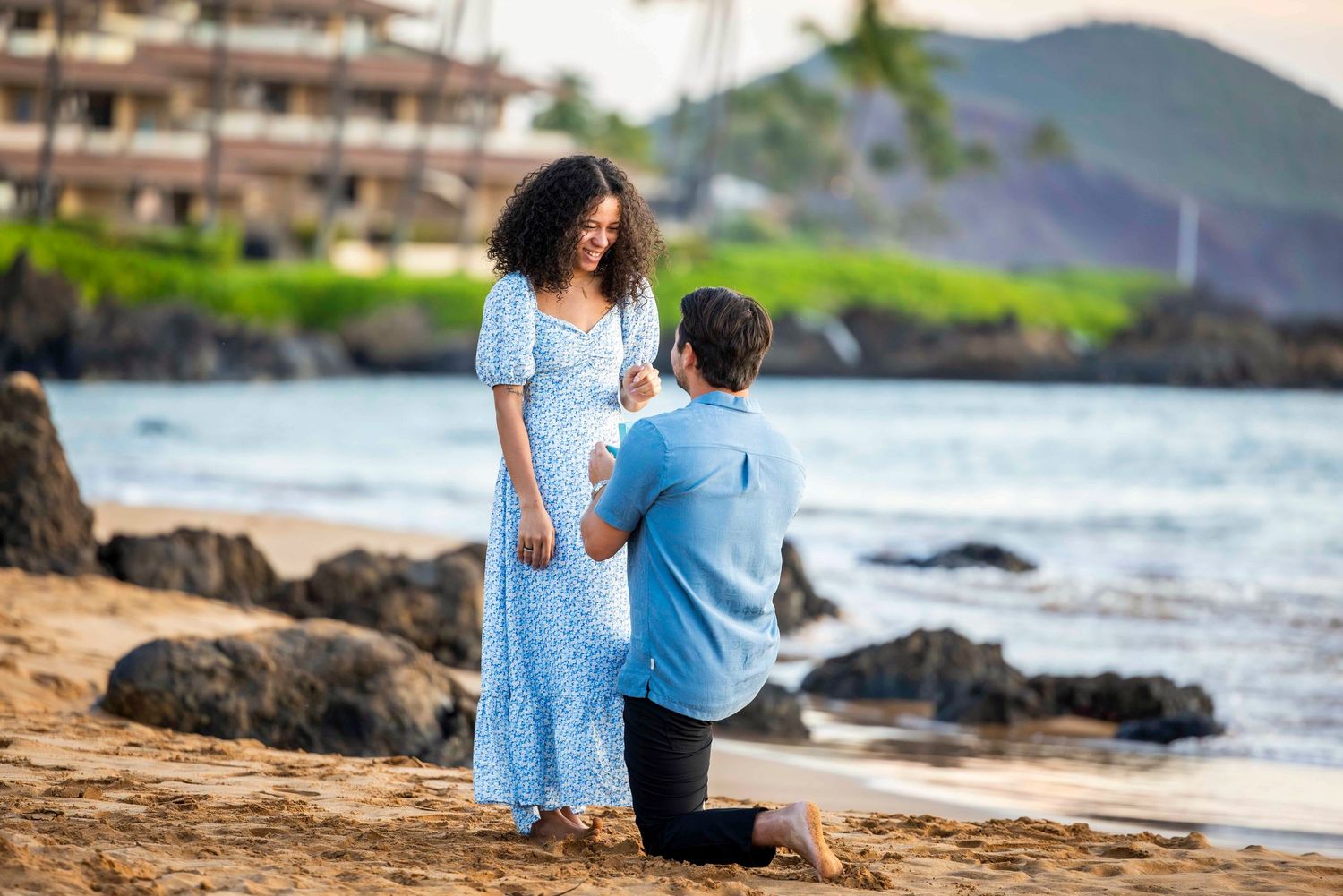 Man in a blue shirt kneels on a sandy beach proposing to a smiling woman in a light blue dress, with ocean waves and rocks behind them.