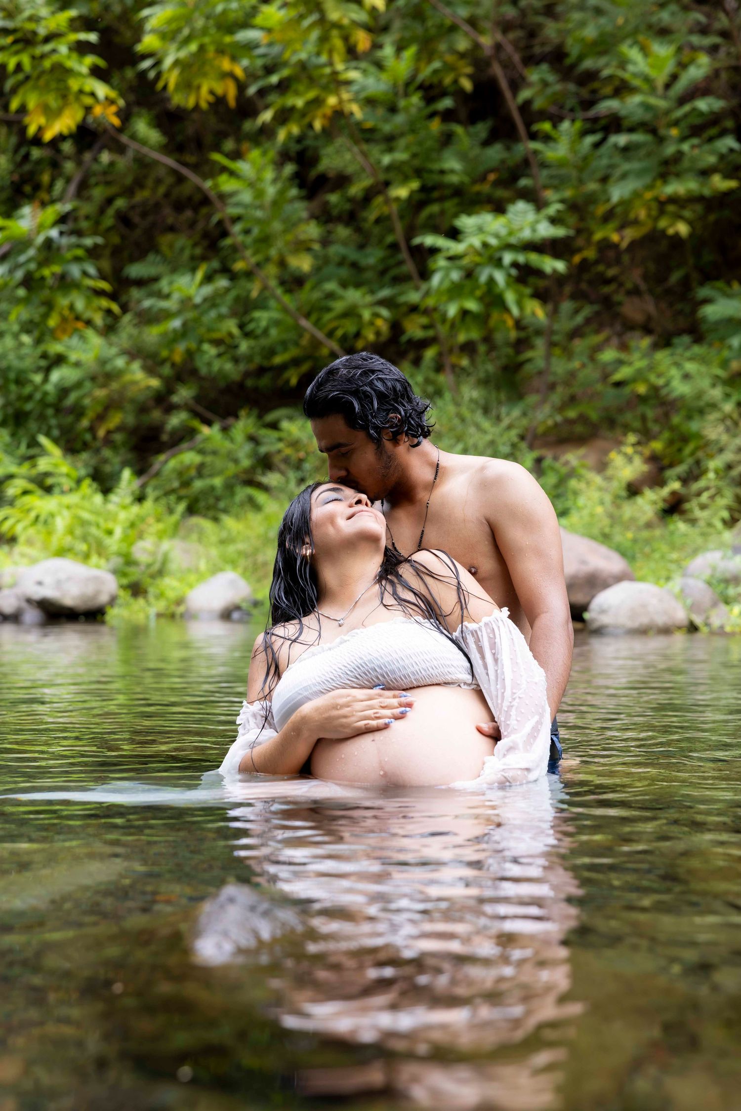 Pregnant woman and her partner embrace in a shallow river surrounded by lush greenery during an outdoor maternity photoshoot. Iao Valley
