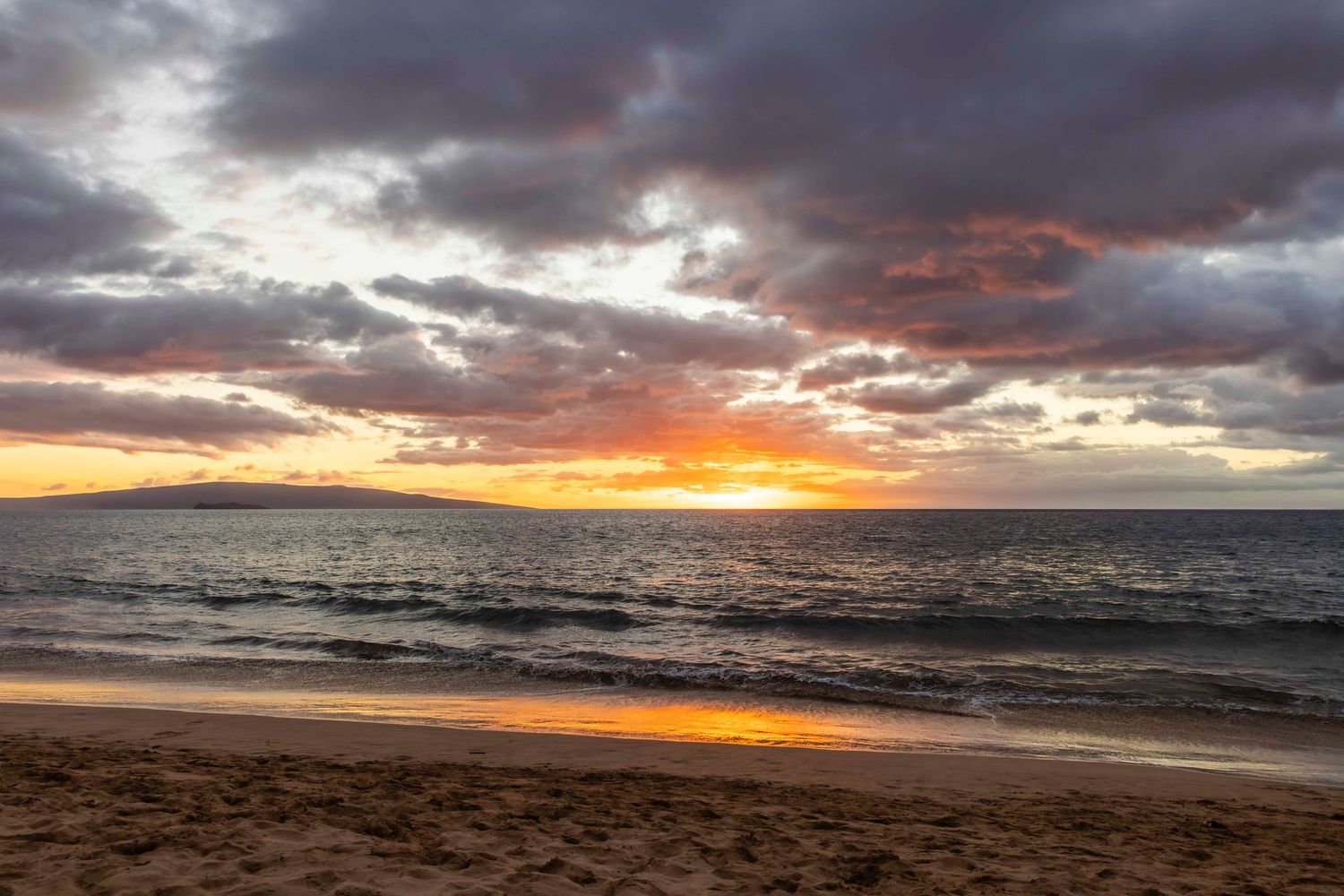 Golden sunset over a calm ocean with dramatic clouds and gentle waves washing onto a sandy tropical beach at dusk.