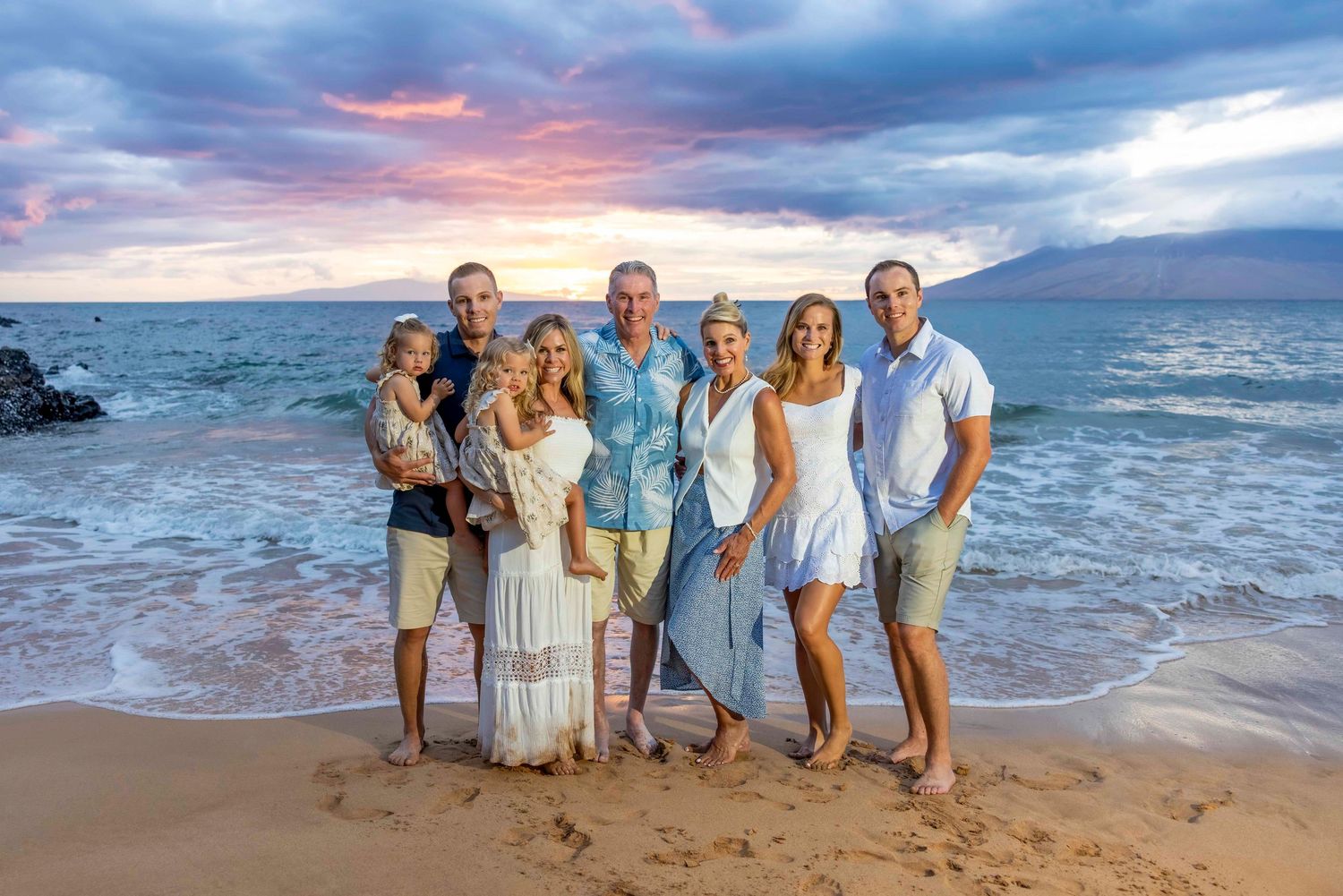Large multigenerational family gathers on a beach at sunset, standing in shallow waves under a colorful pink and purple sky.