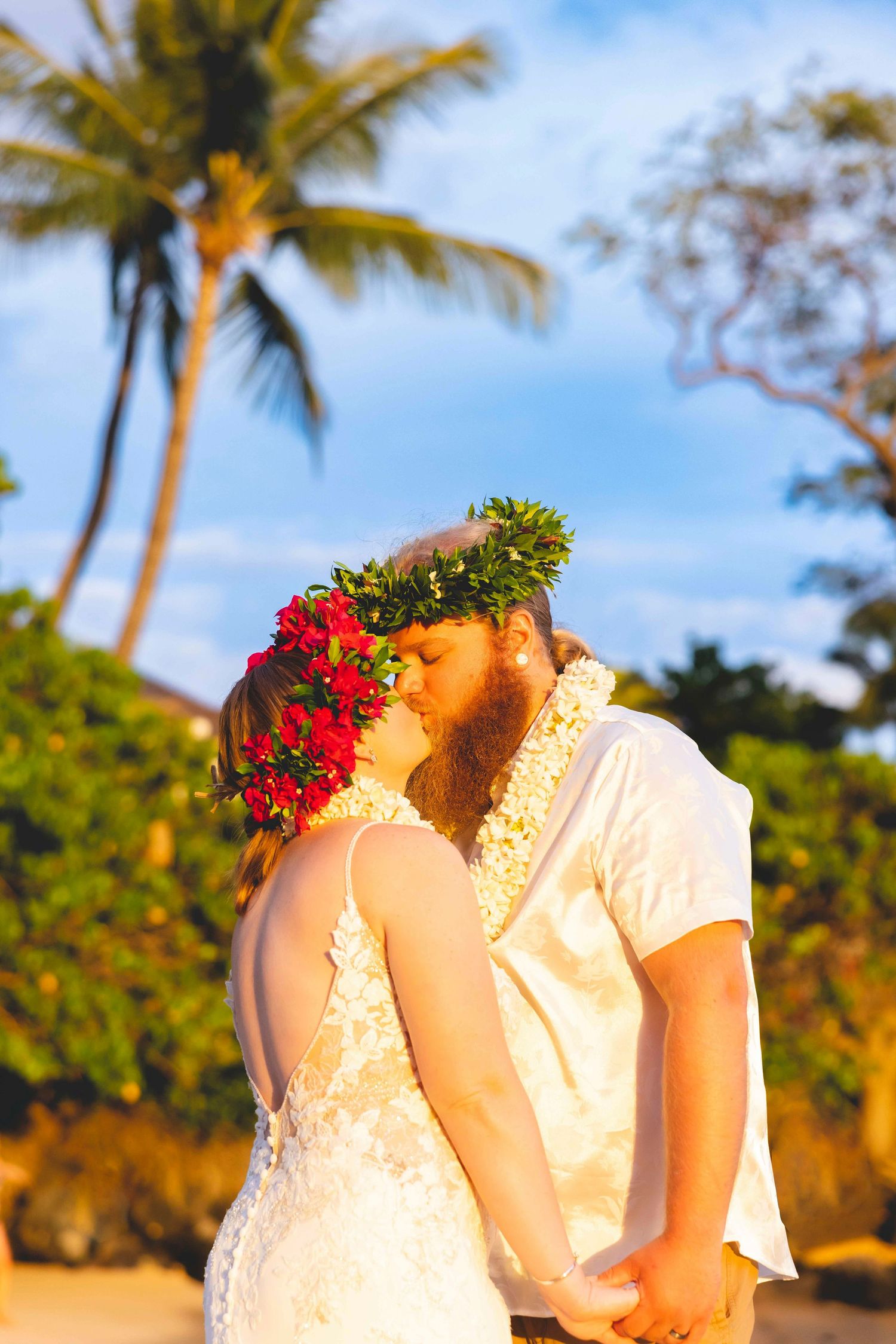 Couple in Hawaiian leis and flower crowns share a kiss on a tropical beach at sunset, the bride in a lace gown beneath palm trees. Makena