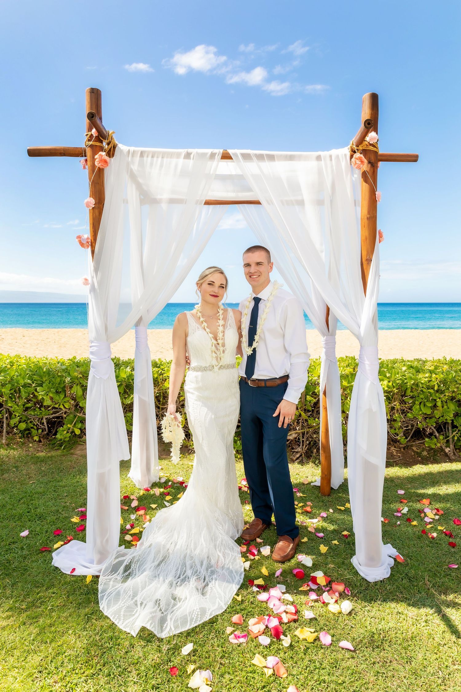 Bride and groom pose under a white draped wooden wedding arch with flowers on a tropical beach lawn, rose petals scattered on the grass.