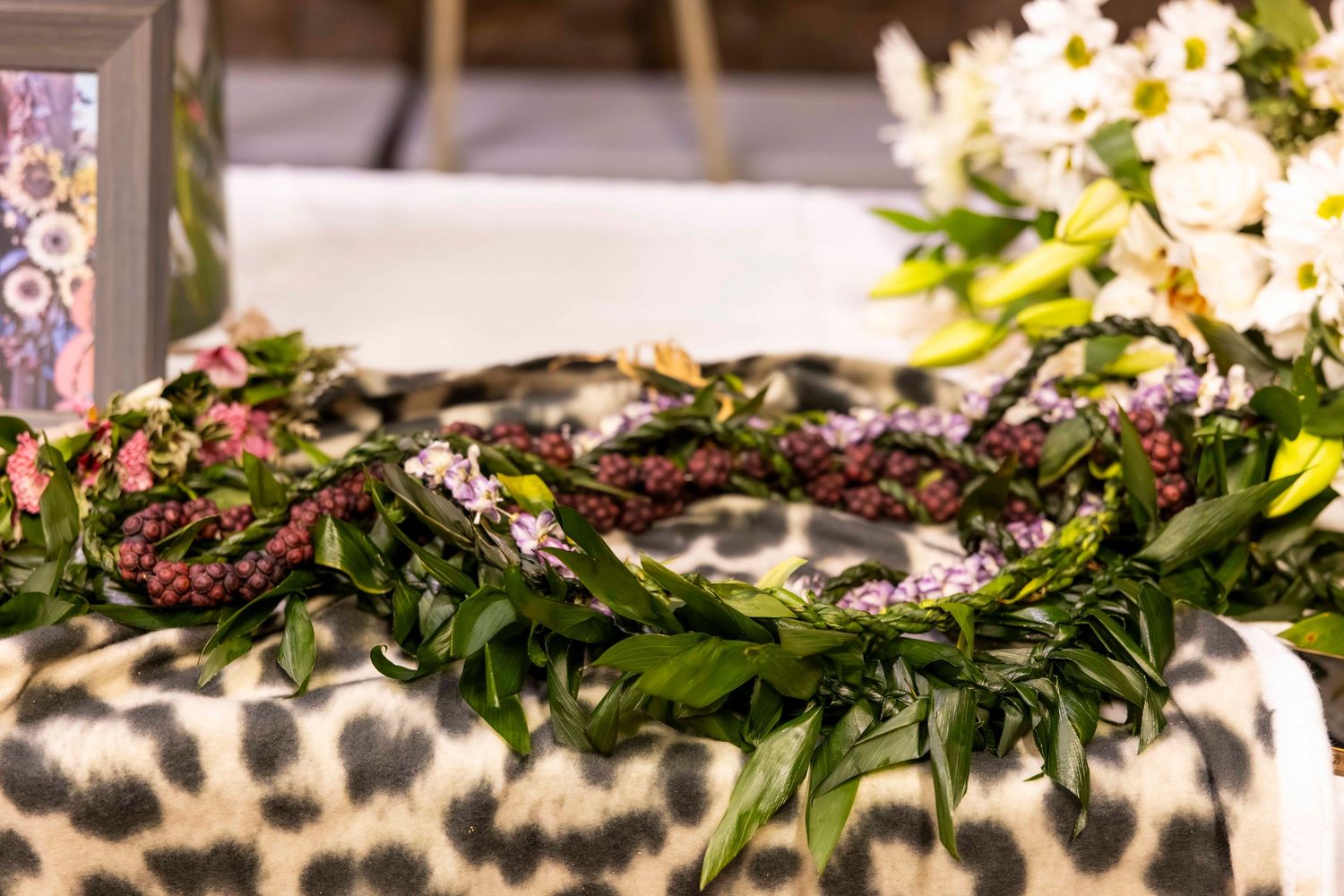 Traditional Hawaiian maile and purple flower leis with green leaves draped over leopard print fabric beside white blossoms.