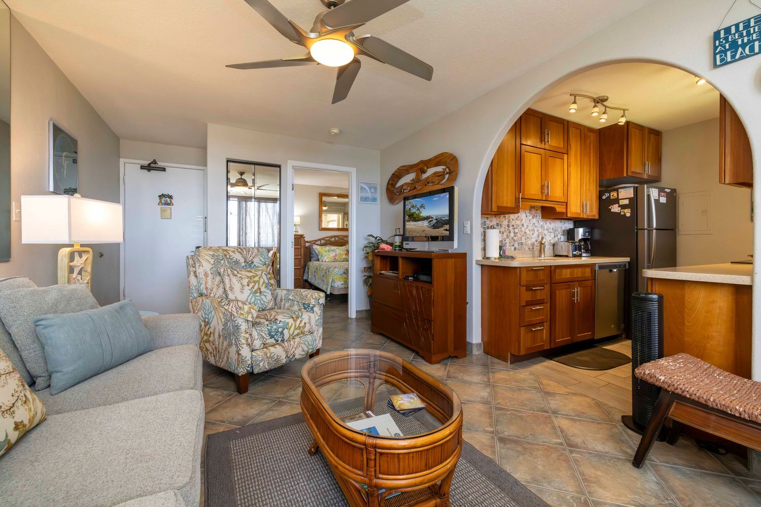 Cozy coastal condo living area with gray sofa, floral armchair, oval rattan coffee table, ceiling fan, and open kitchen with wood cabinets.