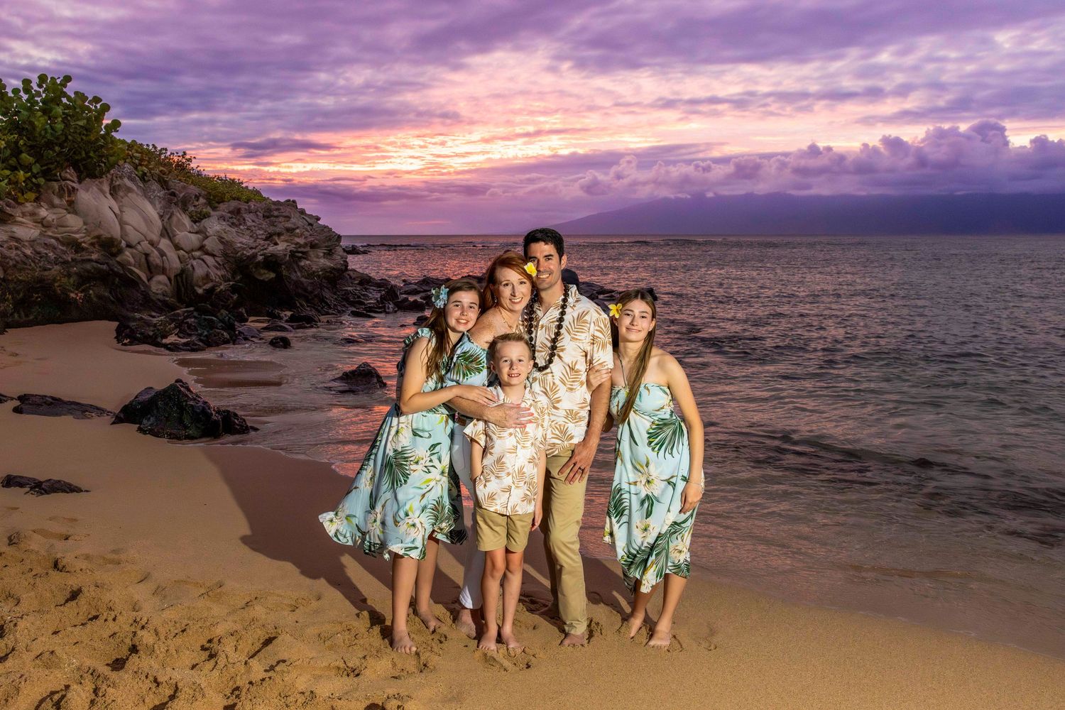 Family of five in tropical print outfits stands on a sandy beach at dusk with a purple and pink sunset sky over the ocean.