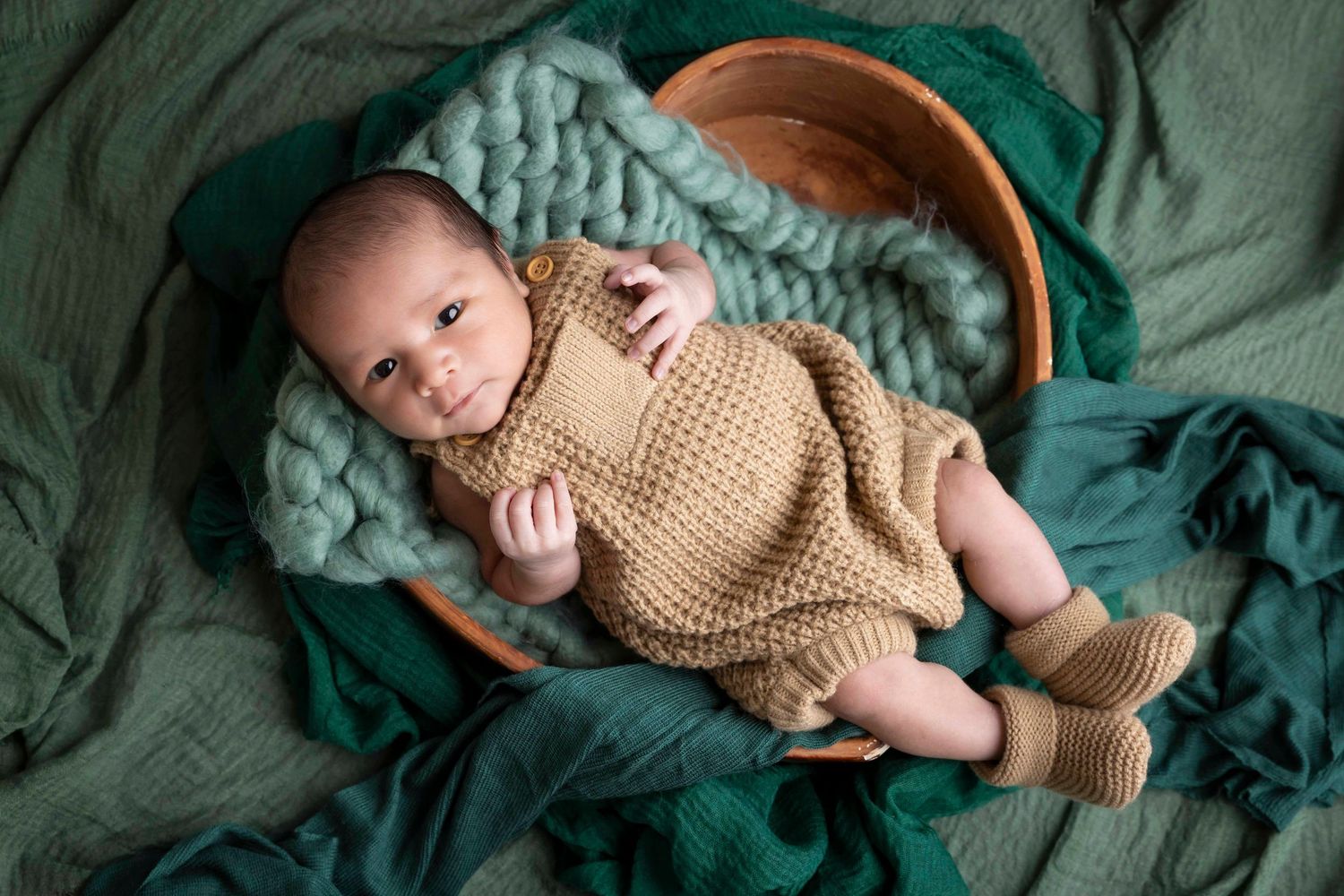 Baby in a knit tan romper and booties lying on green blankets and chunky wool for a cozy newborn portrait.