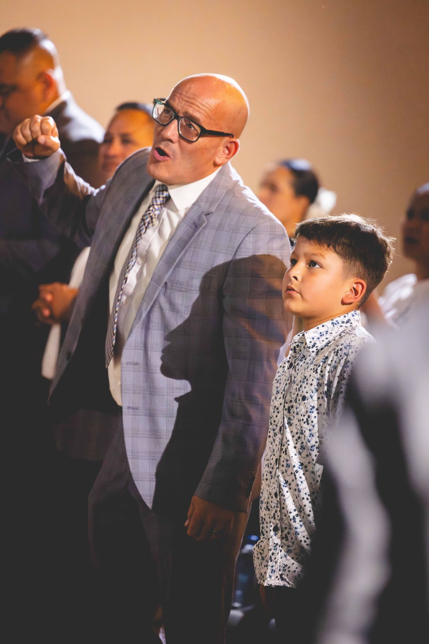 A bald man in glasses and a plaid blazer raises his fist while a young boy in a patterned shirt stands beside him in a crowd.